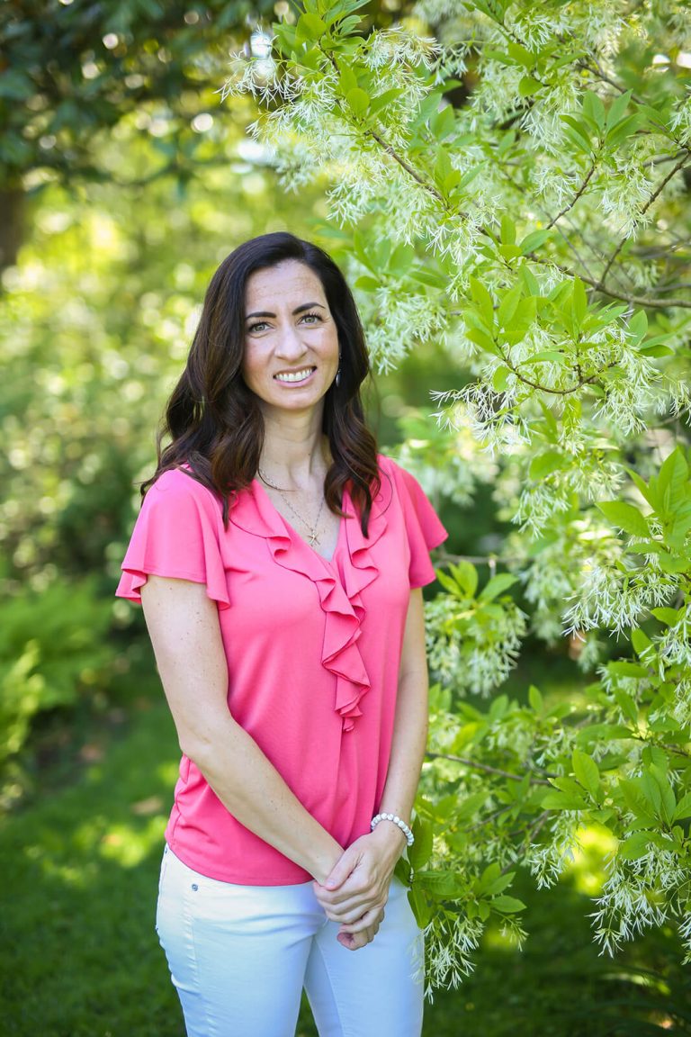 Erin Shepherd, SEO and content strategy consultant for small business owners, wearing a pink shirt and white pants standing in front of a tree with white flowers.